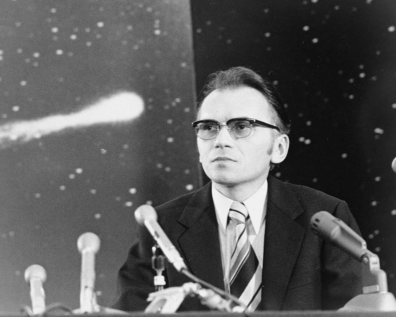 Photograph of Kohoutek seated behind several microphones