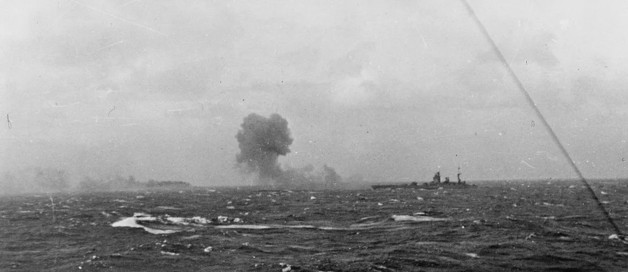 Rodney firing on Bismarck, which can be seen burning in the distance