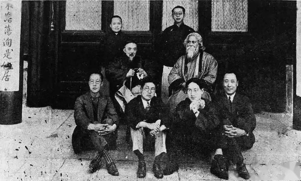 Posed group black-and-white photograph of seven Chinese men, possibly academics, in formal wear: two wear European-style suits, the five others wear Chinese traditional dress; four of the seven sit on the floor in the foreground; another sits on a chair behind them at centre-left; two others stand in the background. They surround an eighth man who is robed, bearded, and sitting in a chair placed at centre-left. Four elegant windows are behind them in a line.